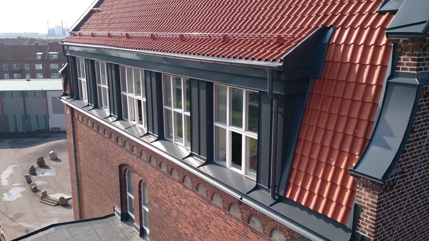 Takkupa i mörk plåt längs tegelfasadens fönsterrad. Bild 1 av 5