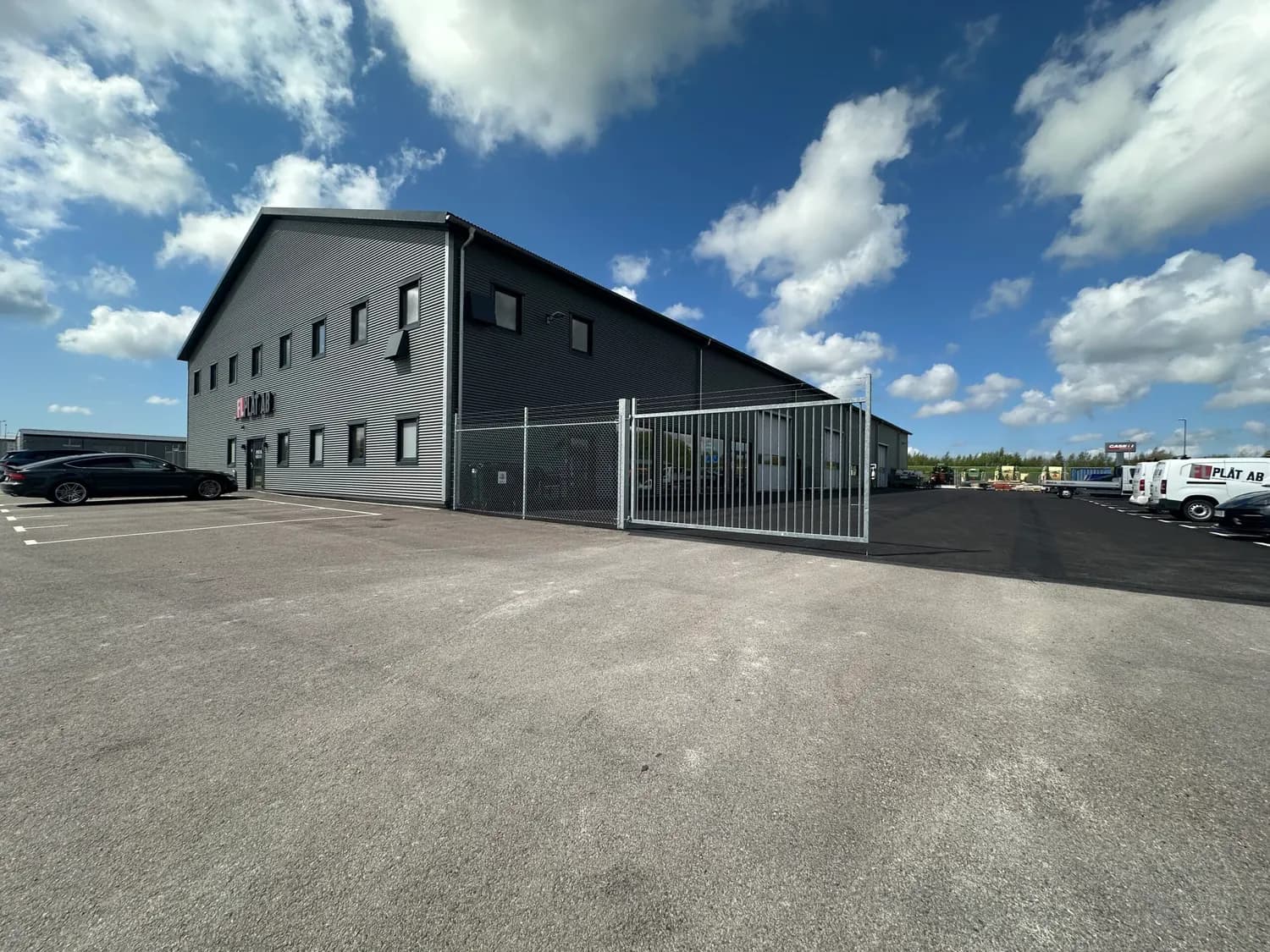 Hela byggnaden utifrån en molnig dag, med grind och parkering. Bild 16 av 16