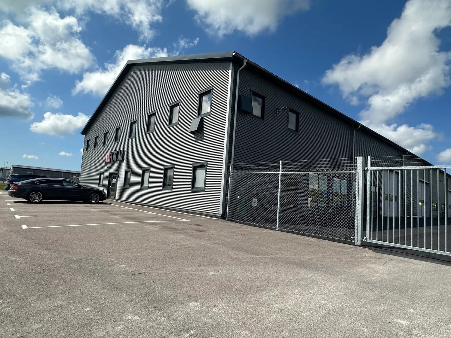 Byggnaden snett från hörnet, mörk plåtfasad och grind mot gården. Bild 3 av 16