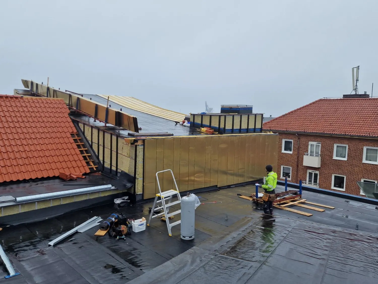 Kopparfasaden möter grannhusets tegeltak, arbete pågår på taket. Bild 3 av 21