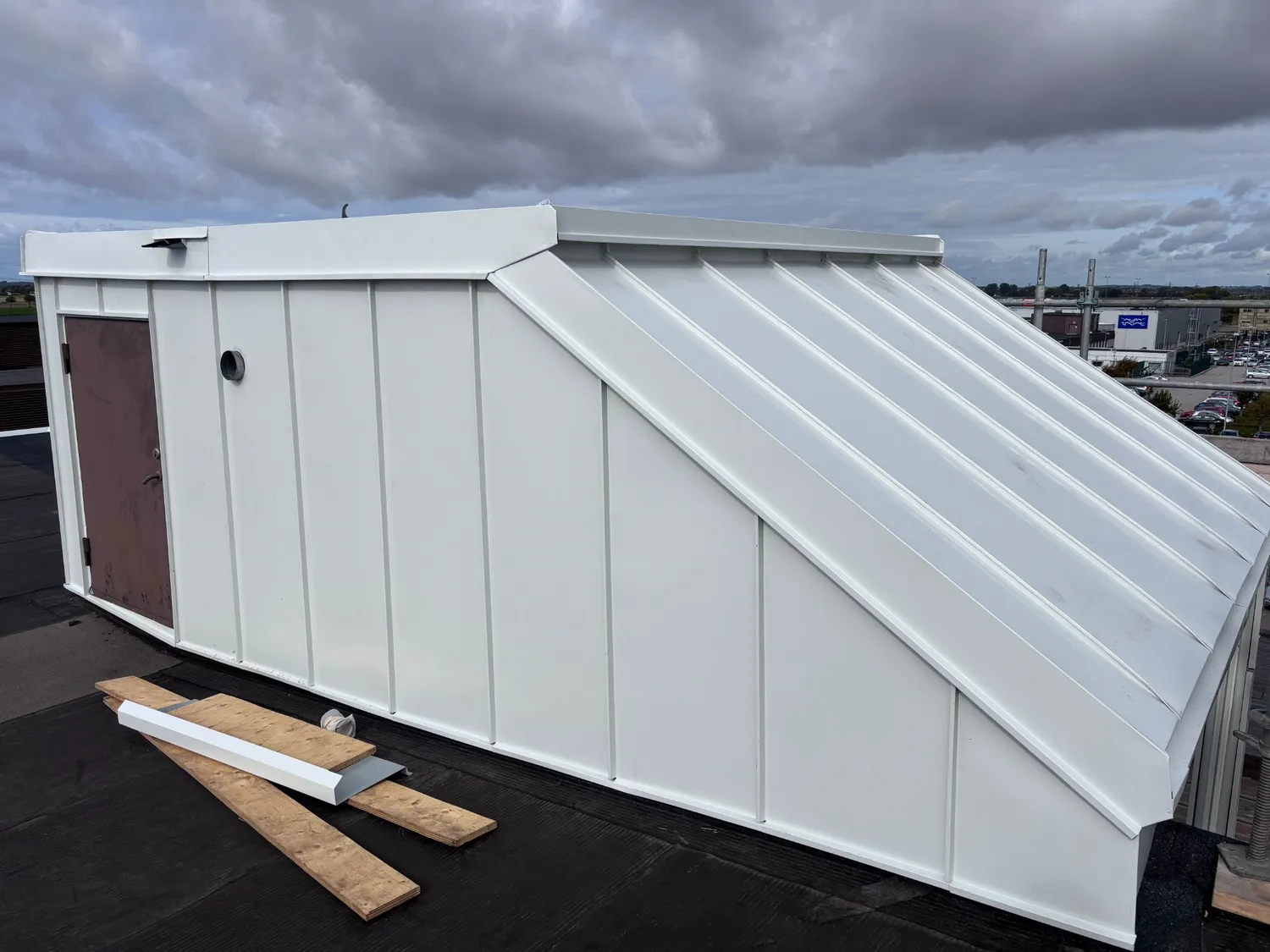Takhus klätt i vit stående fals med bruna dörren och ett runt ventilationsgaller, virke och plåtprofiler på takduken i förgrunden. Bild 6 av 11