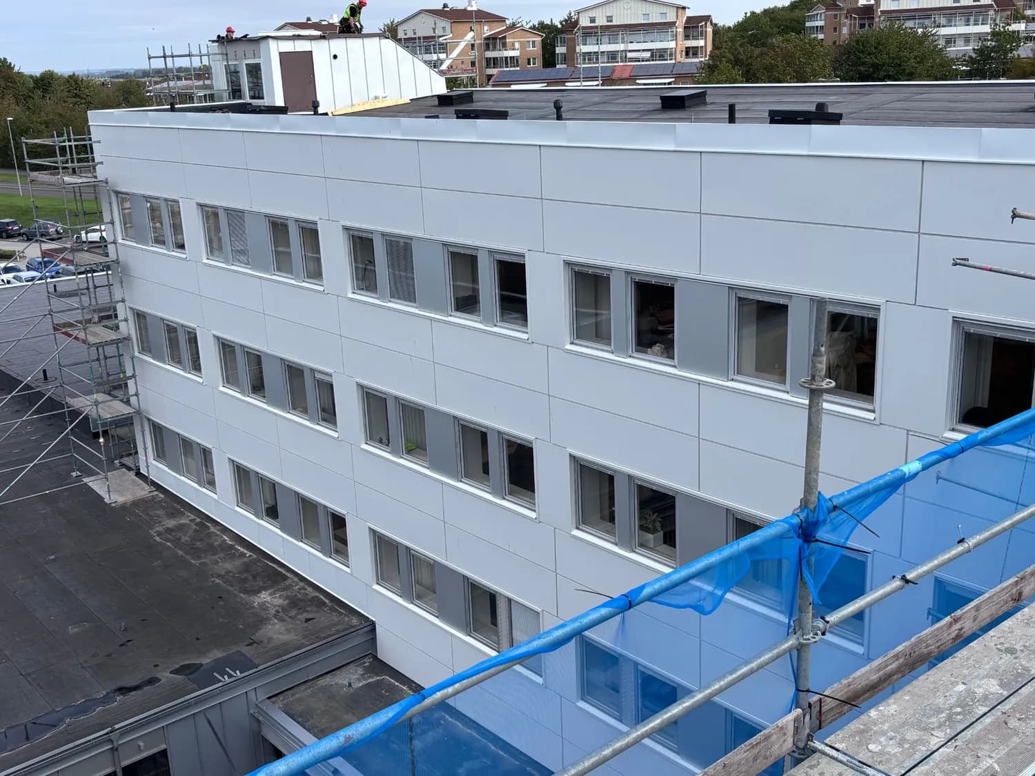 Vy från ett angränsande tak ner mot fasaden med vita fasadkassetter och fönsterband i fyra våningar, ställning till vänster och blått skyddsnät till höger. Bild 7 av 11