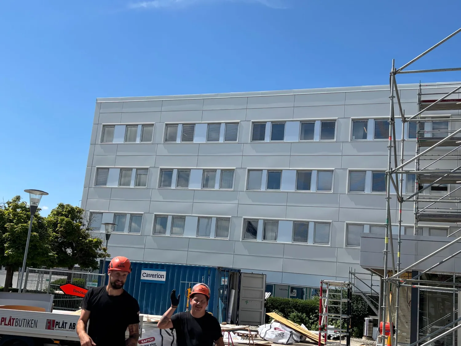 Två plåtslagare i hjälm framför den färdiga vita fasaden en solig dag, med FL Plåts bil och ställning i bakgrunden. Bild 8 av 11