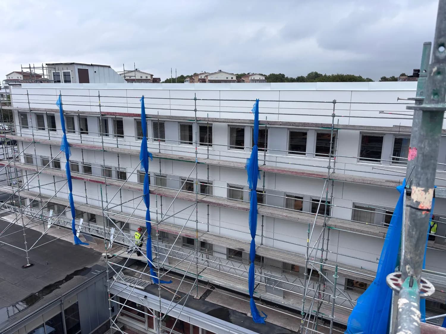 Fasaden bakom ställningens blå skyddsnät, med en byggnadsarbetare i varselväst på en av plattformarna. Bild 9 av 11
