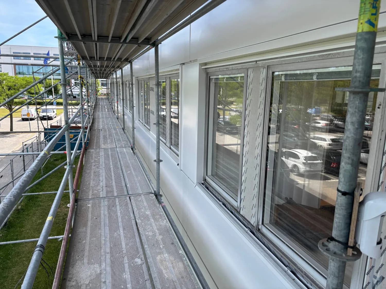 Gångbrygga i ställningen längs fasaden, vita fasadkassetter monterade kring fönstren med skyddstejpen kvar på karmarna. Bild 10 av 11