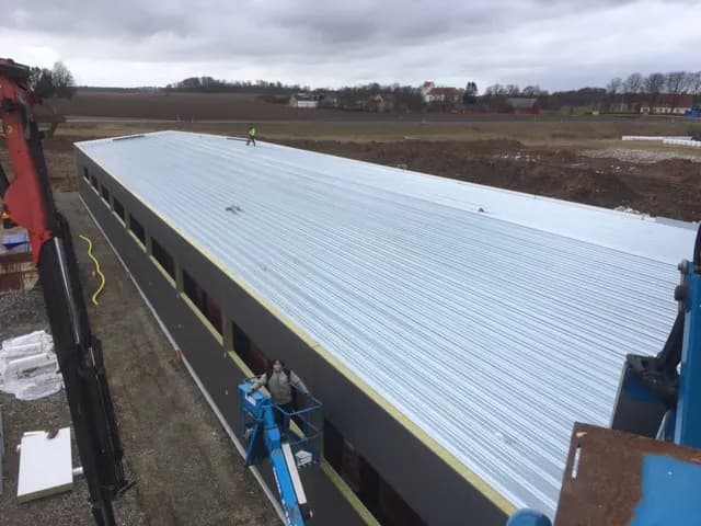Takplåten lagd över hela hallen, plåtslagare i skylift nedanför. Bild 19 av 23