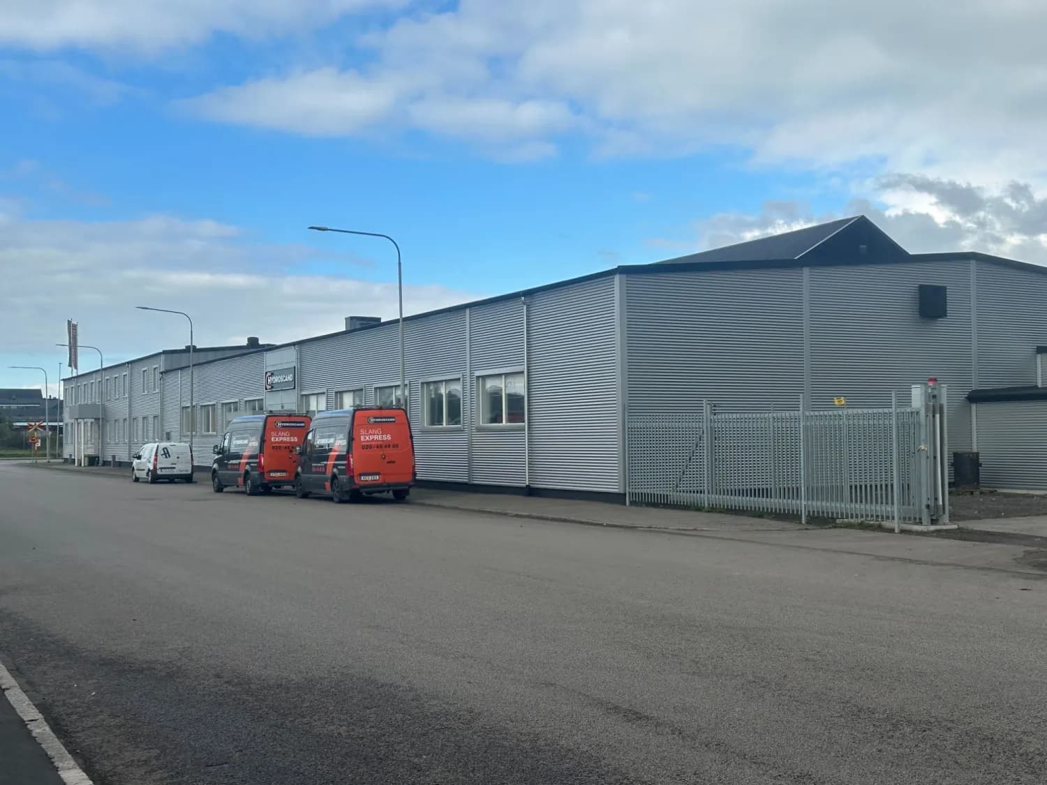 Efter: verkstadshallen längs gatan i samma ljusgrå vågprofilerade plåt, med fönsterband och servicebilar parkerade utanför. Bild 4 av 4