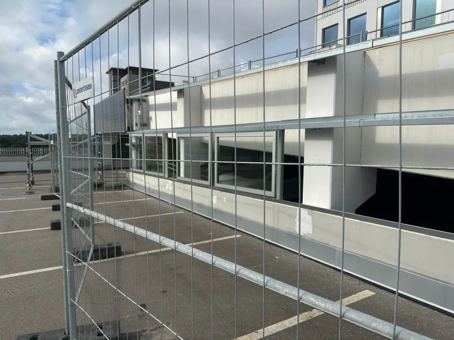 Före: den gamla betongväggen på parkeringsdäcket, sedd genom byggstängslet, med ett nytt sockelbeslag i aluminium längs foten. Bild 1 av 4
