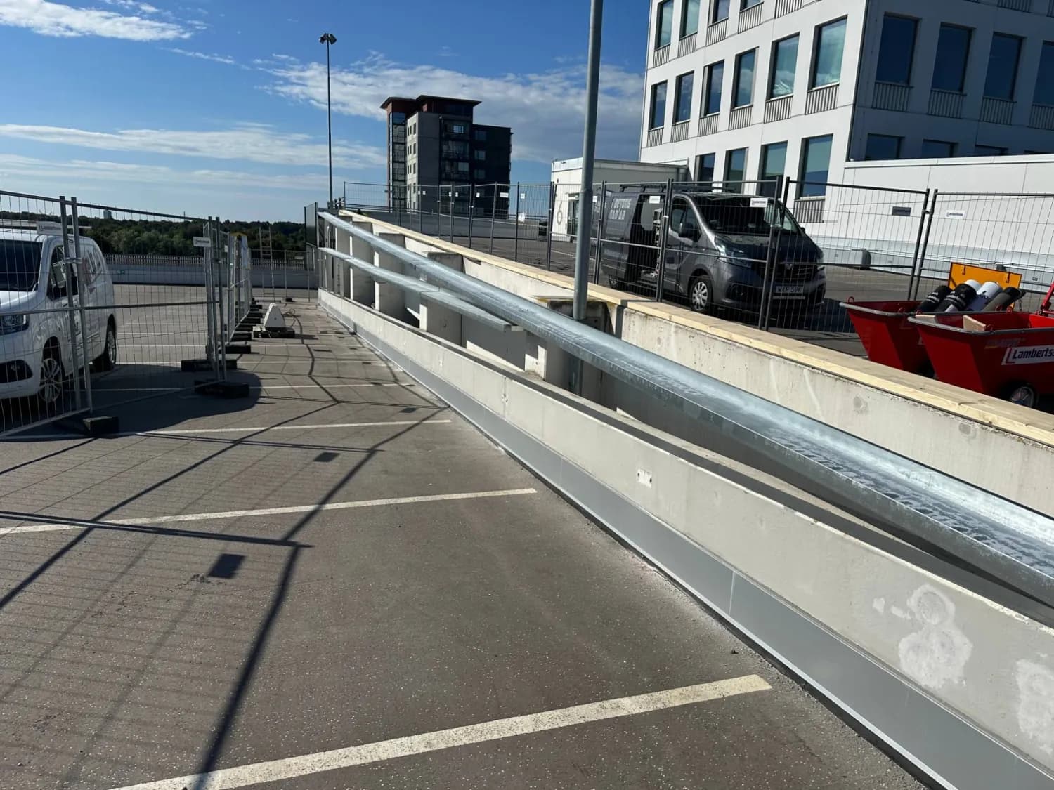 Galvaniserade profiler utlagda längs muren inför krönbeslaget, med färdigt sockelbeslag i aluminium vid foten och byggstängsel runt arbetsplatsen. Bild 3 av 4