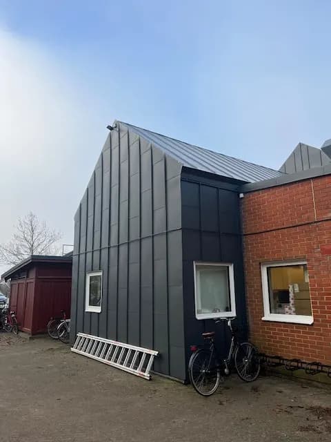 Tillbyggnad klädd i mörk plåt intill den äldre tegelbyggnaden. Bild 4 av 9