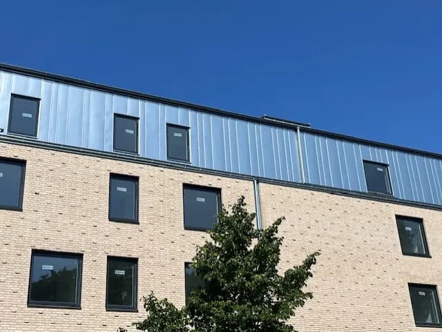 Färdig fasad i ljust tegel med plåtklädd överdel och träd framför. Bild 12 av 13
