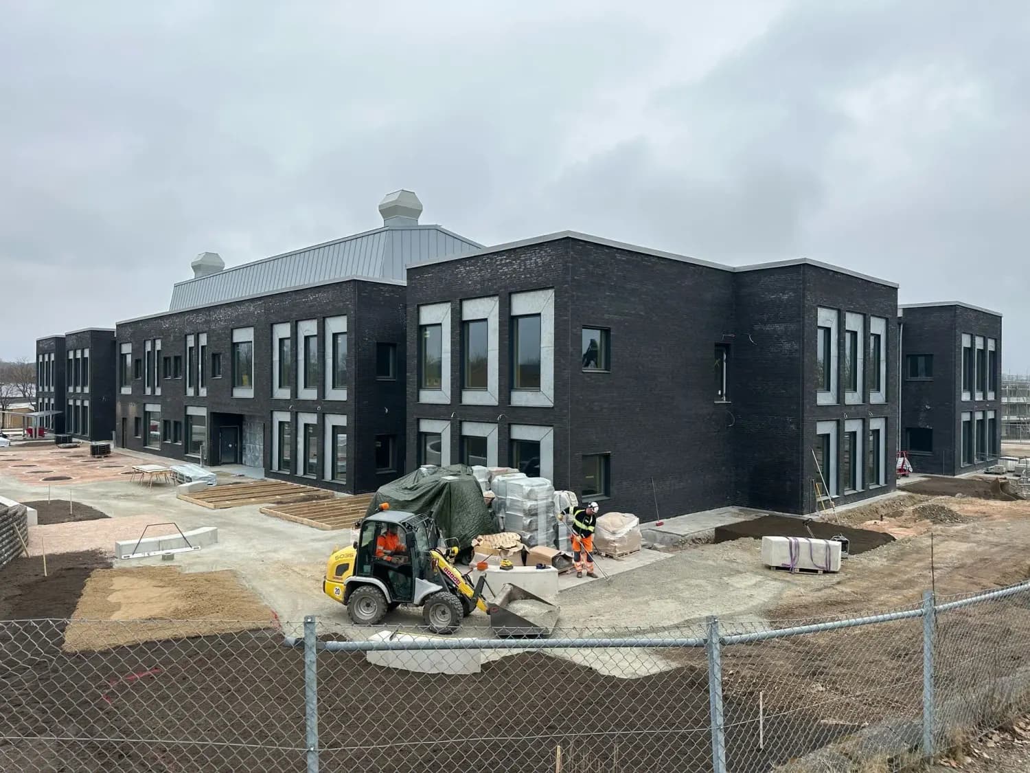Byggnaden i sen fas med hjullastare och materialpallar på gården. Bild 12 av 19