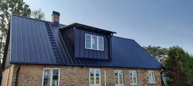 Takkupa i mörk plåt med spröjsat fönster över tegelfasad. Bild 60 av 62