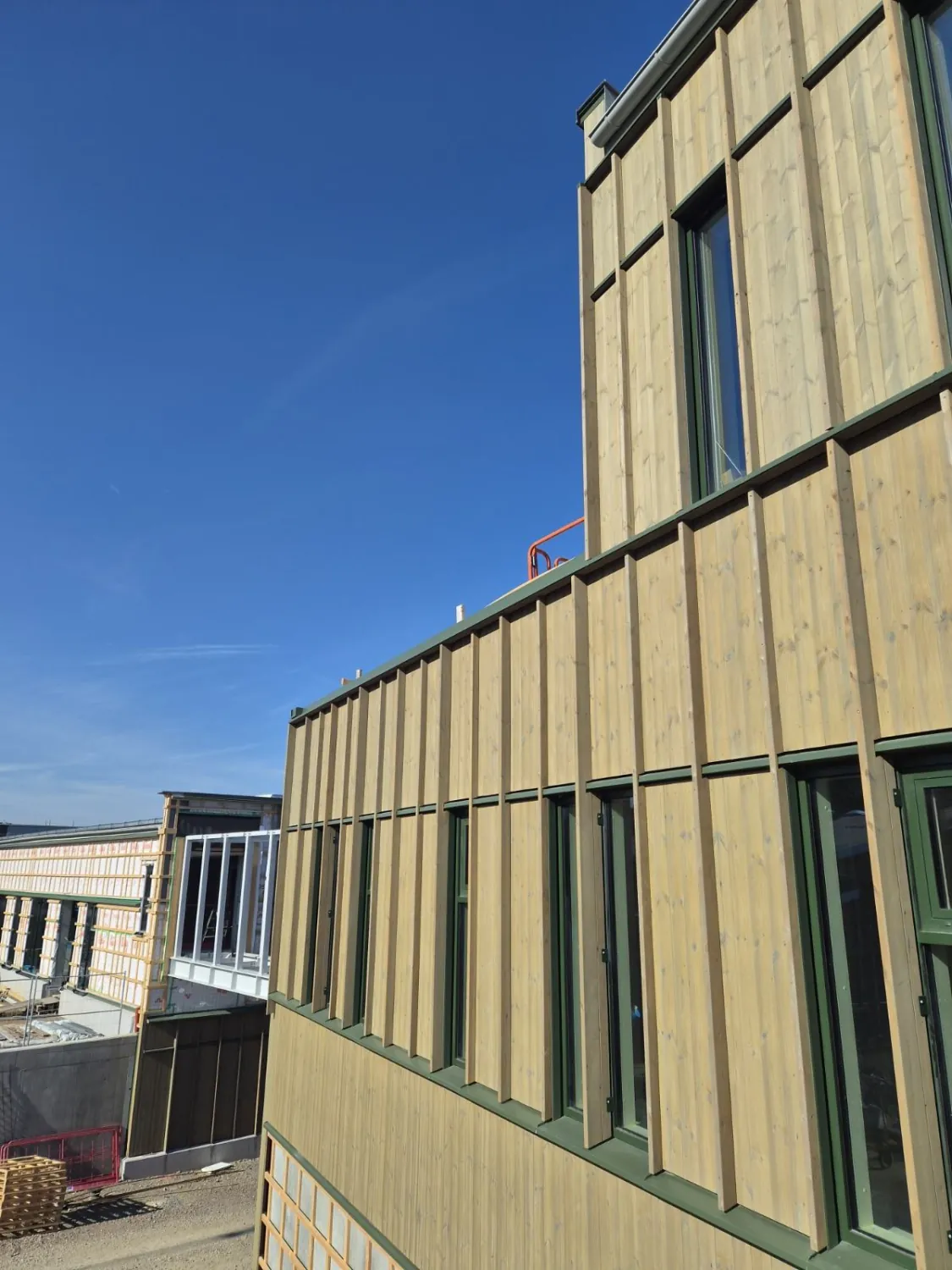 Mossgröna droppbleck i varje skarv mellan panelfälten på två våningar av träfasaden, med en byggnadsdel i vindskydd i bakgrunden. Bild 9 av 12