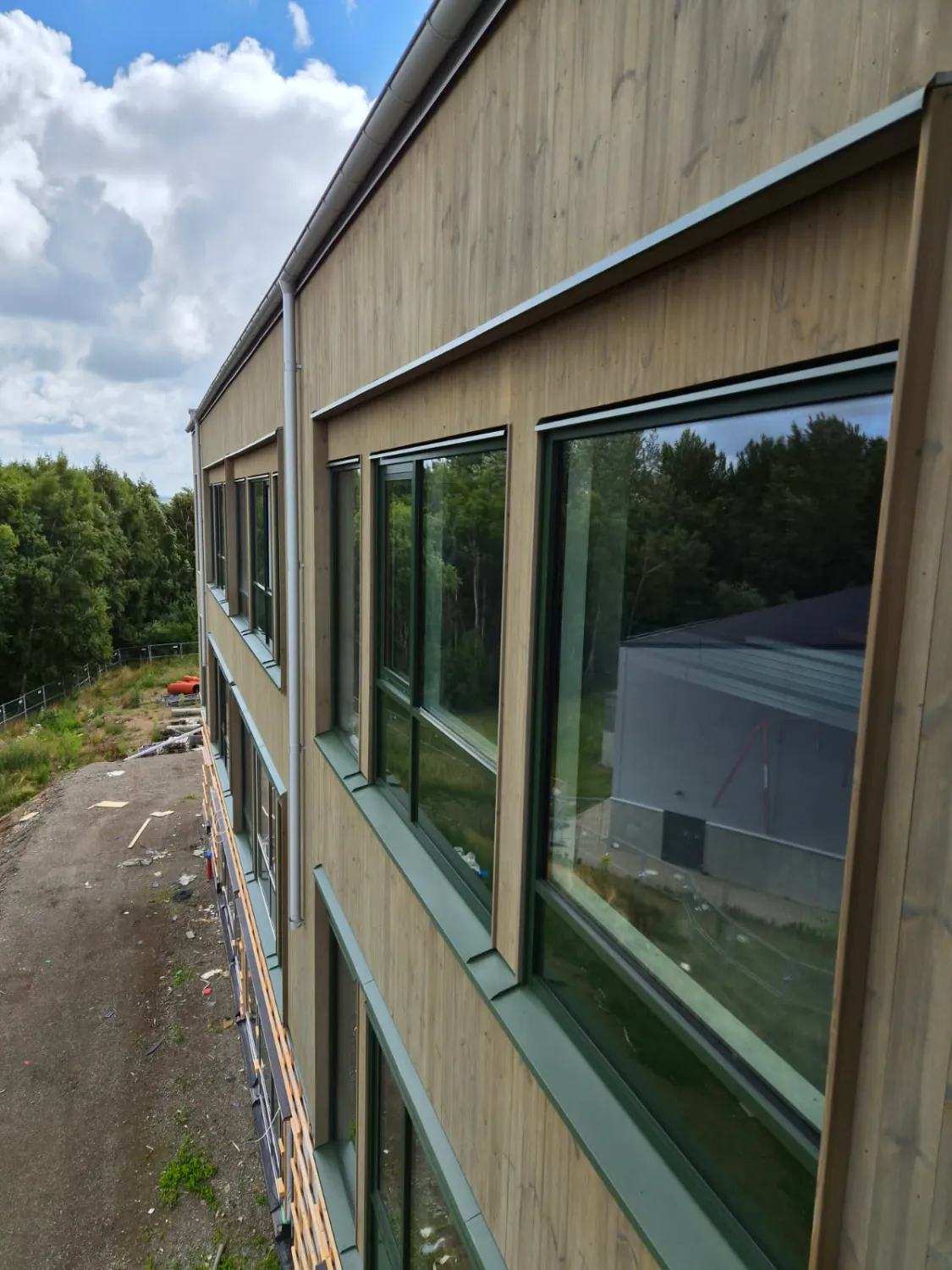 Fönster i träfasaden inramade av överbleck och fönsterbleck i mossgrön plåt, med stuprör och hängränna i ljusgrått. Bild 8 av 12