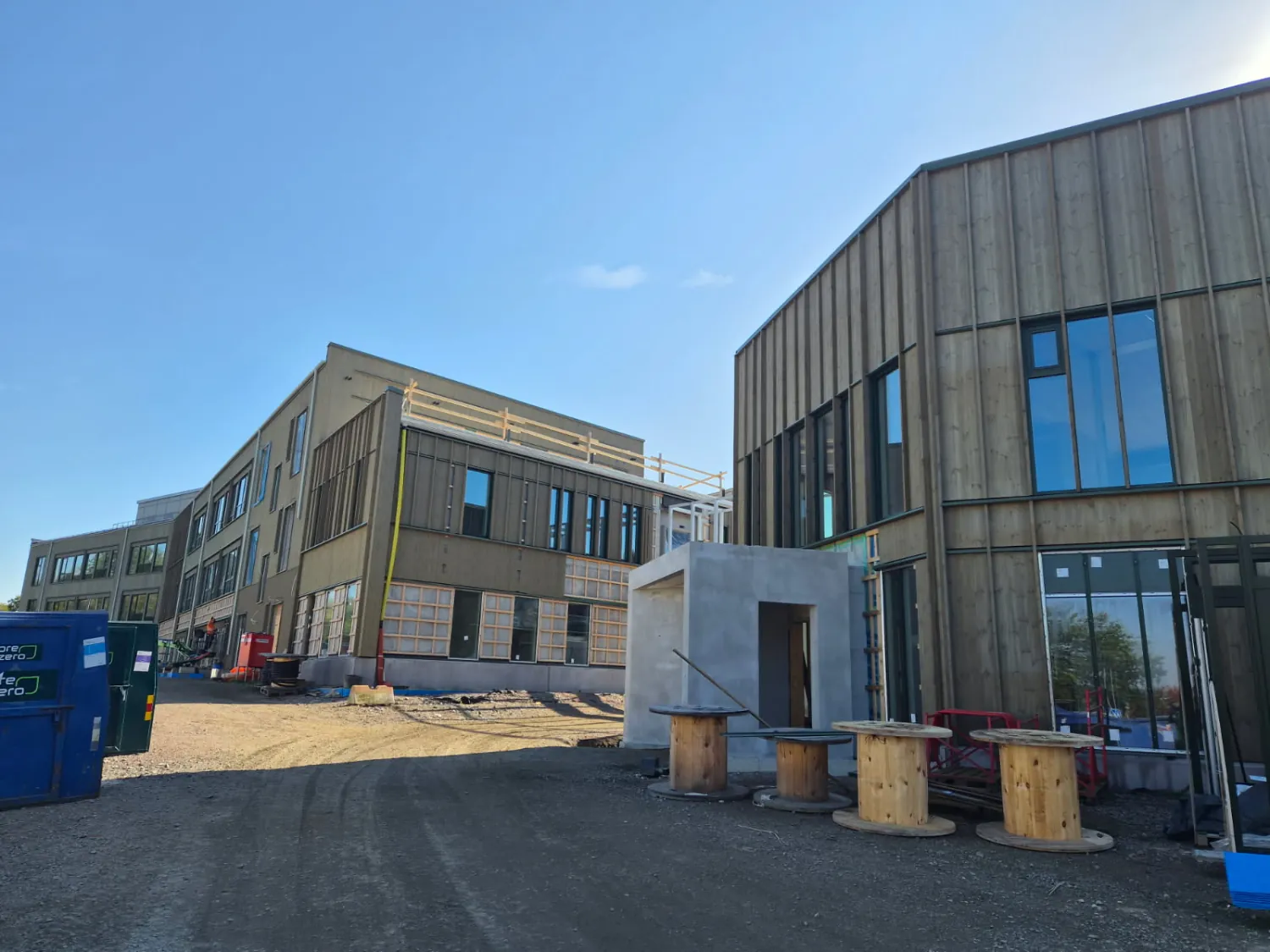 Skolbyggnaderna sedda från gården, med lodrät träpanel och plåtbeslag i skarvarna mellan våningarna, kabeltrummor och containrar i förgrunden. Bild 2 av 12