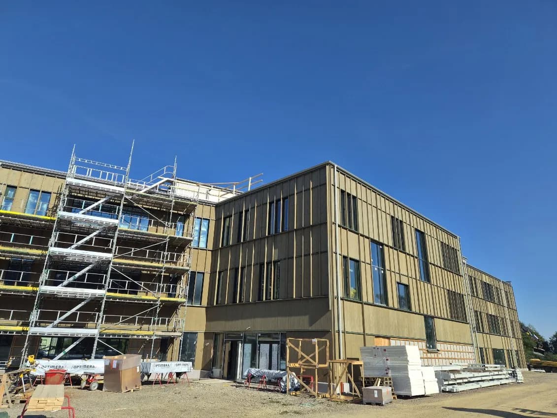 Skolans tre våningar med träfasad och plåtbeslag längs fönsterrader och takfot, med ställningen kvar vid ena gaveln och byggmaterial på gården. Bild 1 av 12