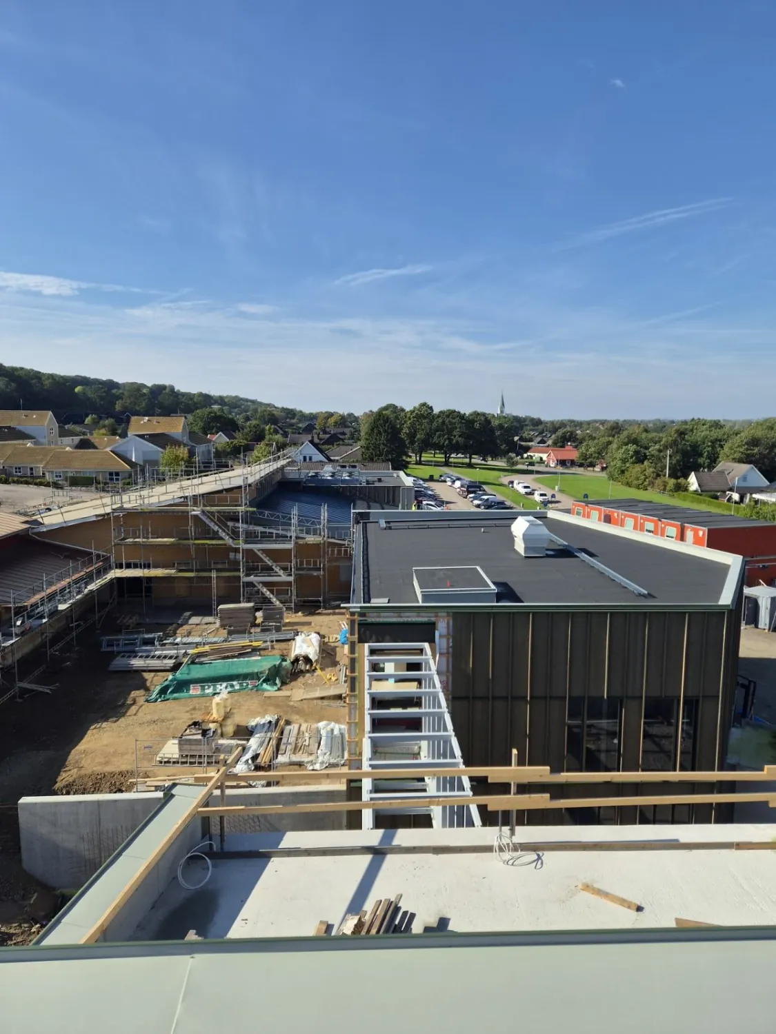 Utsikt från taket över skolbygget, med ljusgrå krönbeslag i förgrunden, en byggnadsdel i ställning till vänster och villorna och ett kyrktorn i bakgrunden. Bild 12 av 12