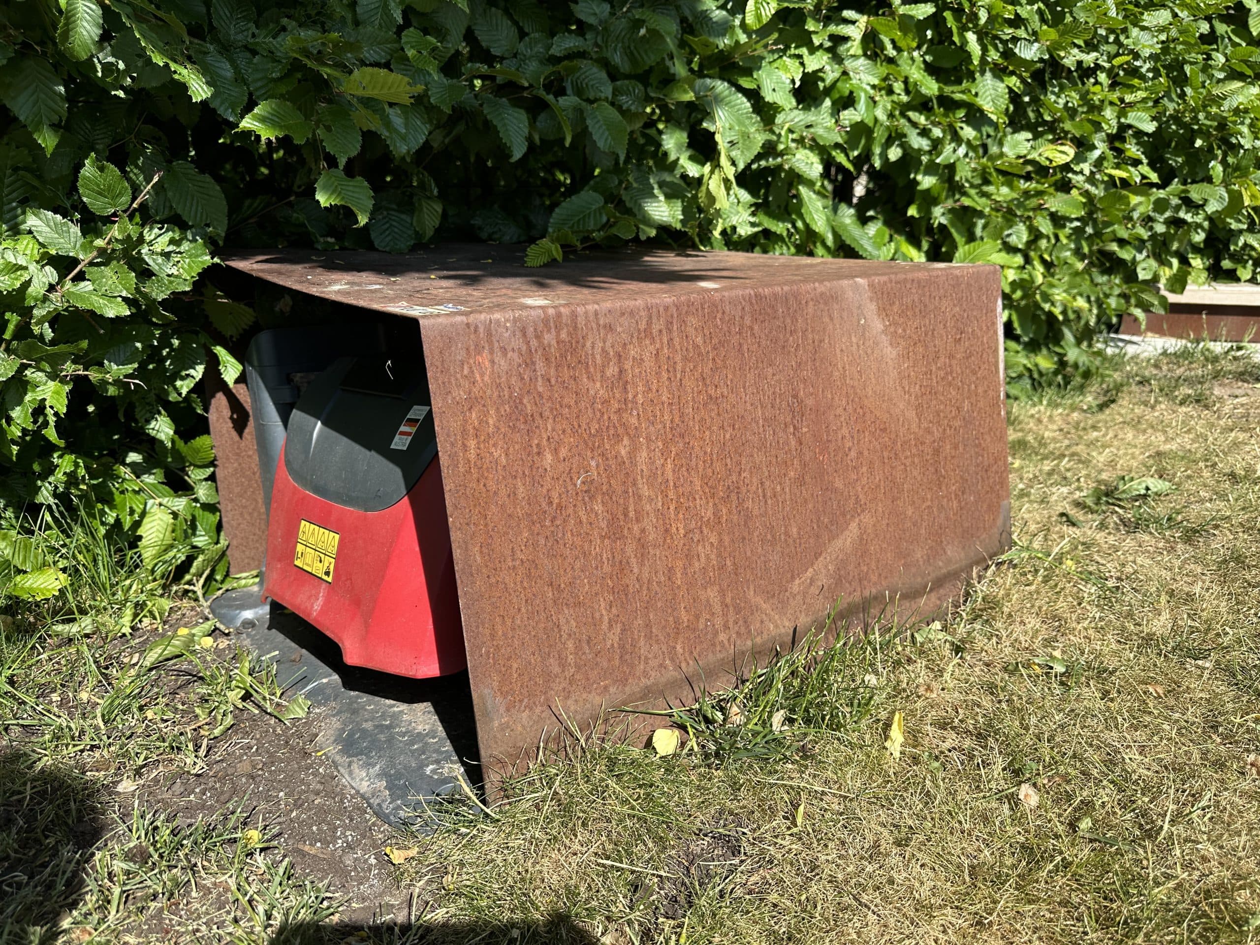 Hus i cortenplåt över en robotgräsklippare vid en häck
